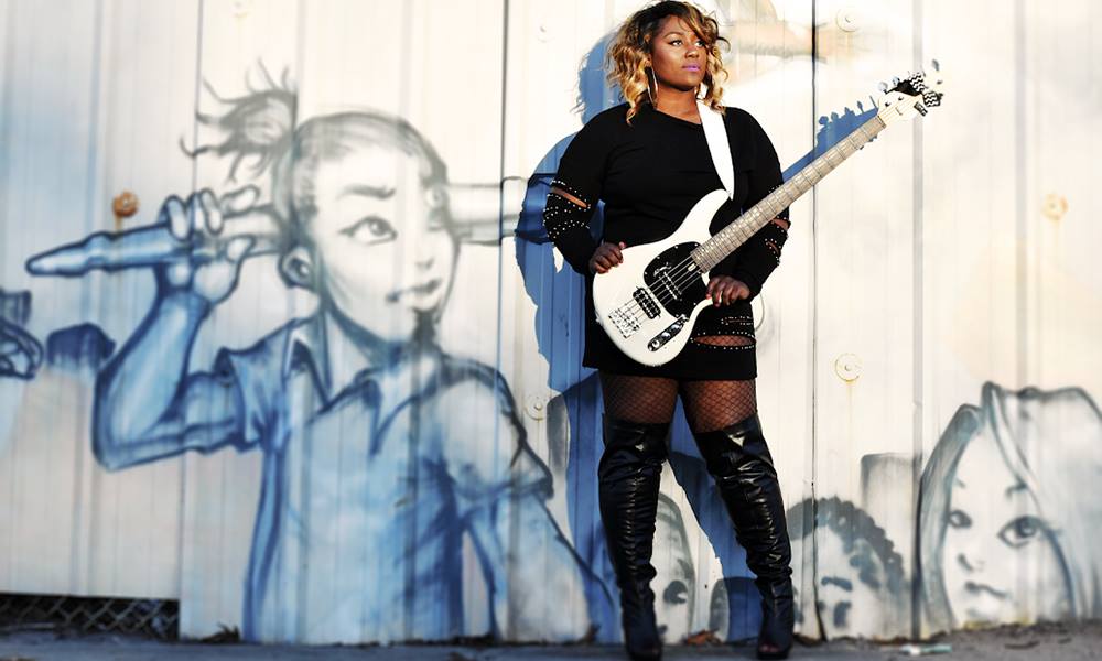 Teja Veal with her bass in front of a fence featuring graffiti image of a girl.